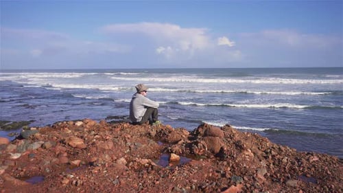Mann mit Hut meditiert am roten Felsstrand im sonnigen Sommer, Natur, Ozeanküste