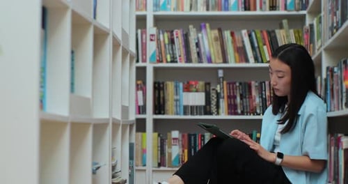 Asian Girl Sitting on the Floor Near Shelves with Book in the Library Uses Digital Tablet Computer