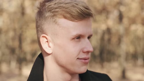 Portrait d'un bel homme en manteau noir assis près de l'arbre dans la forêt d'automne par une journée ensoleillée