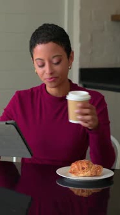 Vertical video: Picking up tablet, woman reading in kitchen with croissant, sipping cup, maroon top