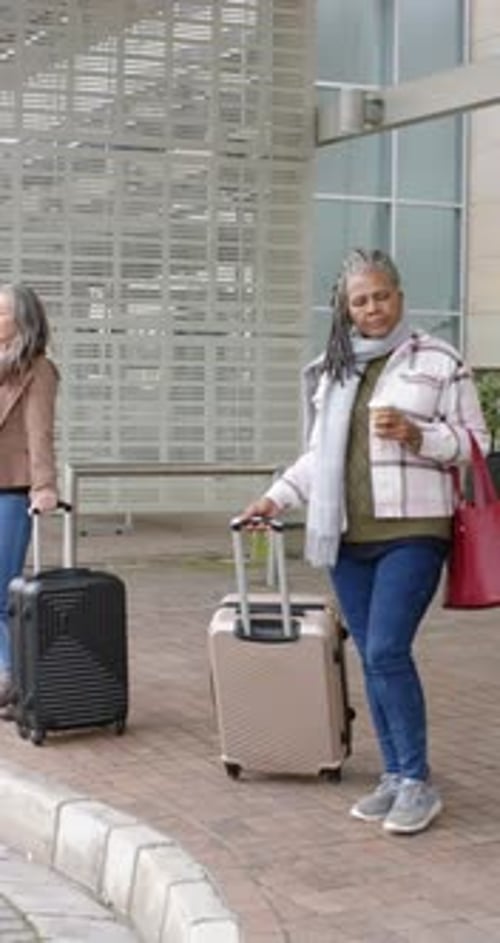 Vertical video: Diverse female travelers at drop-off waiting holding coffee cup pulling suitcases