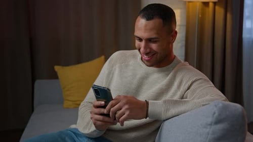 Man Using Smartphone, Smiling on Couch at Home