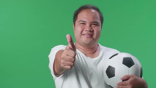 Close Up Of Asian Man With A Ball Showing Thumbs Up Gesture While Cheering Soccer On Green Screen