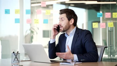 Angry dissatisfied businessman in suit arguing talking on phone sitting at workplace in business