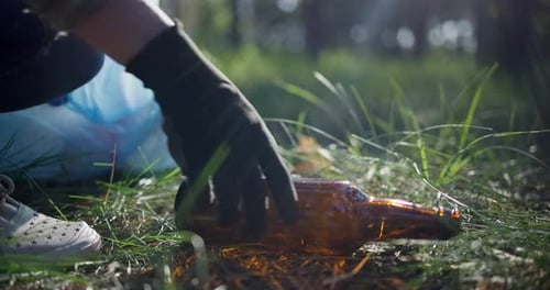 Una mujer con una bolsa de plástico recoge una botella de vidrio tendida en el césped mientras recoge basura en el