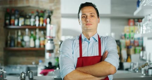 Portrait of a young barman crossing arms and smiling at the camera, behind the bench of a cafe. Co