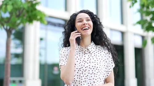 Young smiling female employee is talking on the smartphone while standing near an office building.