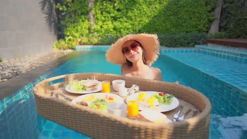 Young Asian woman with a floating exotic breakfast tray in a swimming pool in the hotel resort.