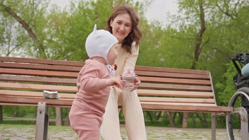 Park Scene With Mother And Baby Mother Sitting On Bench Watching Child Learn To Walk Parent Gently