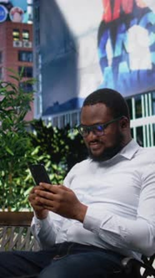 Vertical Video African American Man Sitting on Bench Reading Online Content on His Phone