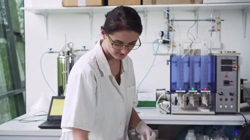 Pretty girl working in laboratory, marking test tube and using laptop, middle shot, front view