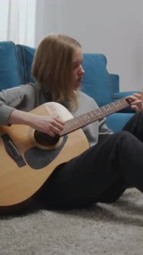 Teen Girl Sitting on Carpet at Home Playing Acoustic Guitar Against Blue Sofa Practicing Thoughtful