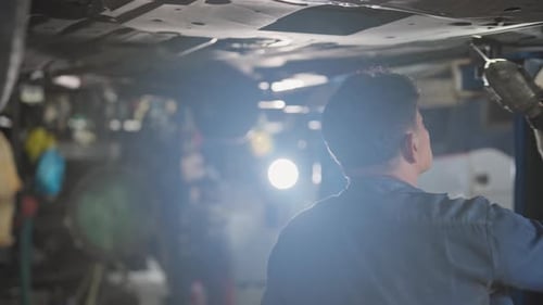 Senior Asian technician or garage worker stand and work under the car and use lantern to guide check