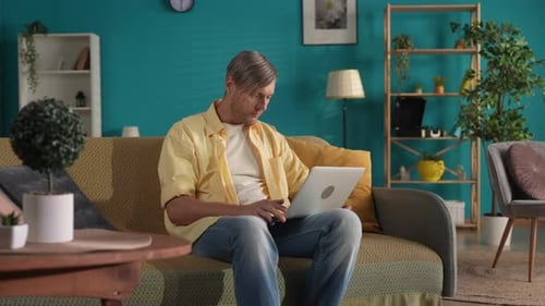 A Man in Casual Clothes Sits on a Sofa in the Living Room with a Laptop on Her Lap A Man Works Types