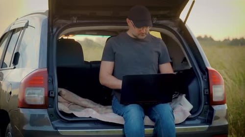 Man Working on Laptop in Car Trunk at Sunset