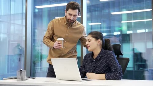 Two colleagues engaged in friendly conversation with coffee at desk at workplace in business office.
