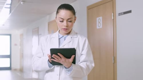 In the Hospital, Beautiful Female Doctor Uses Tablet Computer While Standing in the Hallway. New Mo