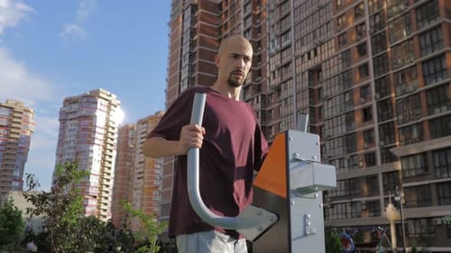 A Young Man with Thin Skin Body is Training on Leg Trainer on a Street Sports Ground in a Beautiful