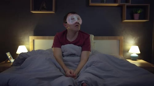 Un homme assis sur un lit soulève un masque de sommeil de ses yeux, se calme et retourne au lit