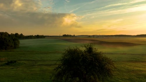 Golden sunlight breaks through wispy clouds over a misty, rolling green field. A wide, atmospheric