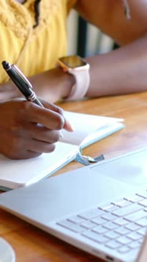 Vertical video: Glancing at laptop, woman in yellow top taking notes in notebook at cafe with pen