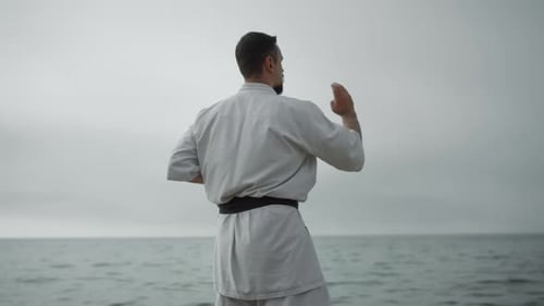 Selbstbewusster Mann übt Handhaltung beim Karate in der Nähe der ruhigen See. Bärtiger Sportler