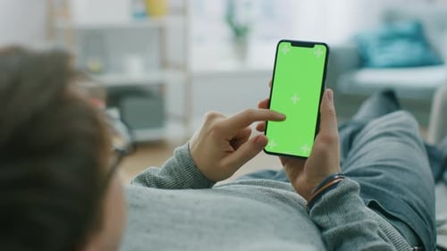 Man at Home Lying on a Couch using Smartphone with Green Mock-up Screen, Doing Swiping, Scrolling G