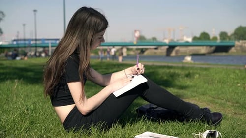 Teenage student writes notes in notebook studying homework in city park