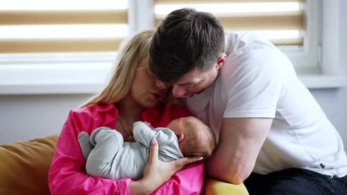 Blonde woman in pink shirt holding tiny newborn. Father is sitting beside soothing his little child.