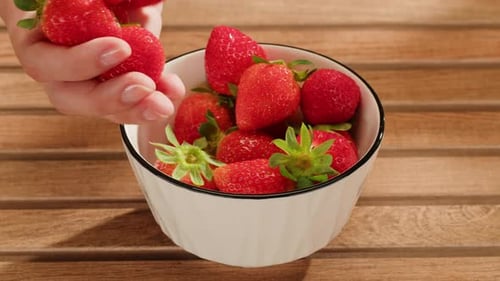 Hand Adding Fresh Strawberries to Bowl on Table