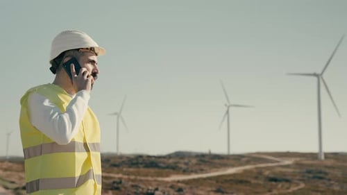 A medium shot of a renewable energy engineer in a field of wind turbines, answering a phone call on