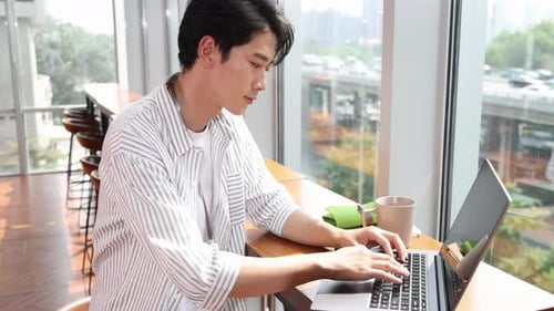 Positive focused busy young business man working sitting by the window typing on laptop computer wor