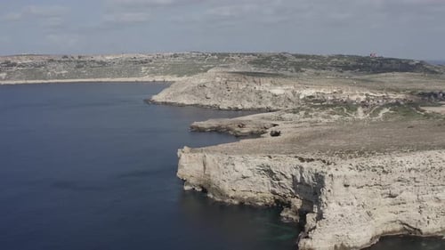 A drone flies backward along Malta’s dramatic dry cliffs, revealing rocky coasts, blue-grey sea, and