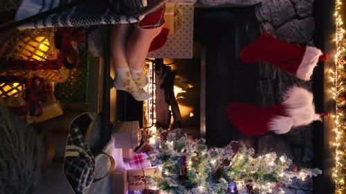Female Feet in Cozy Christmas Woolen Socks Near Fireplace Decorated Xmas Tree