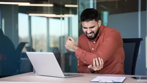 Businessman suffering from elbow pain while sitting at desk at workplace in business office. Upset
