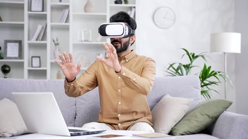 Young bearded freelancer works using VR glasses in virtual reality simulator while sitting on sofa