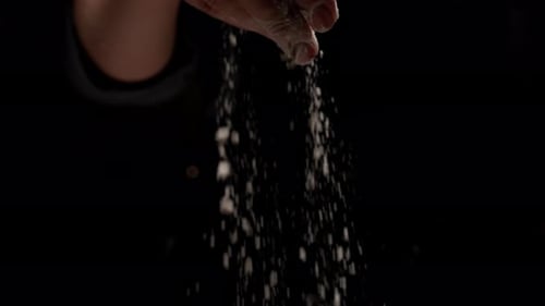 Slow Motion Closeup of a Hand Gracefully Sprinkling Fine White Salt or Flour Against a Dramatic Dark