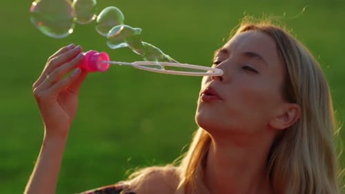 Tender blond woman enjoying summer evening blowing soap bubbles in city park