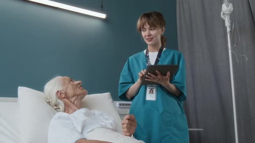 Young Nurse Taking Notes on Tablet Speaking with Senior Patient in Hospital Room