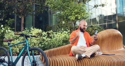 Hombre urbano moderno y hipster en una videollamada en línea con un teléfono inteligente, sentado al aire libre junto a la bicicleta.