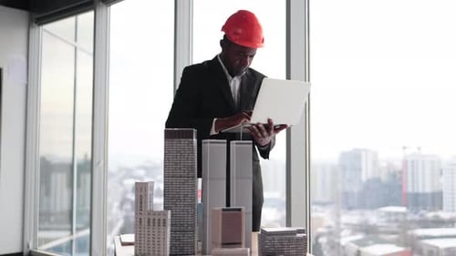 Focused African Man in Suit and Orange Hardhat Using Laptop Looking at Design Megalopolis Model