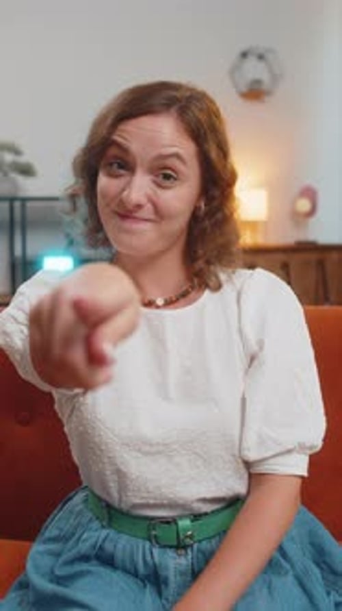 Junge Frau lächelt und zeigt in die Kamera, wählt Einladungen aus und sitzt zu Hause auf der Sofa-Couch