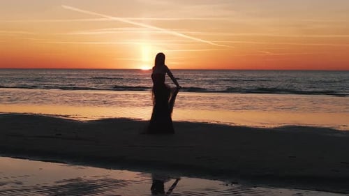 Glückliche schöne Frau in einem langen schwarzen Kleid, das den wunderschönen Sonnenuntergang am Strand in Kalifornien genießt