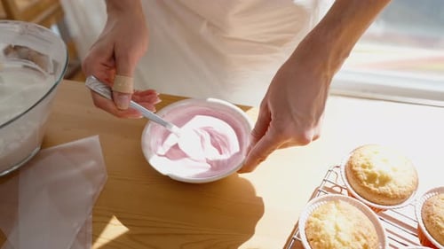 A woman stirs pink frosting for cupcakes in a sunny room. Slow motion.
