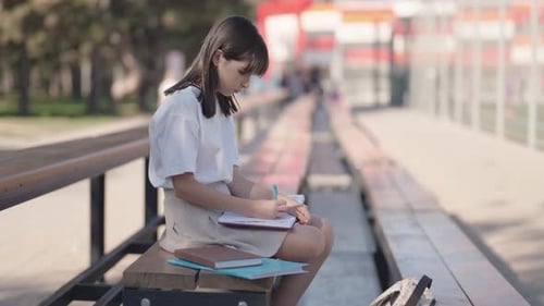 Schoolgirl Sits On Bench After Classes And Writes In Notebook On Sports Ground Near School