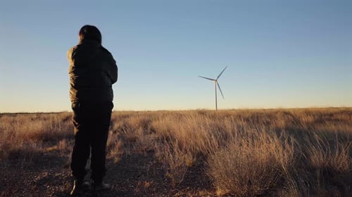 Una persona observa una turbina eólica en una estepa patagónica azotada por el viento mientras los vehículos pasan en la distancia