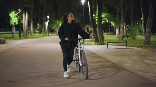 Woman Walking with Bike in Park at Night