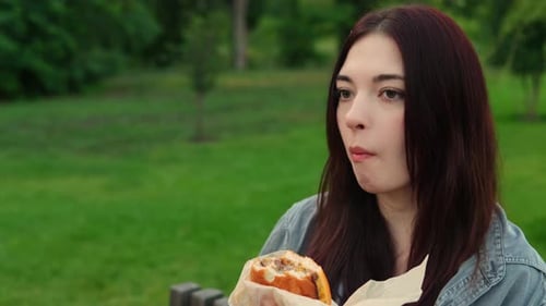 Young Adult Caucasian Lady is Eating Cheeseburger Sitting in the Park Filmed Against Green Lawn