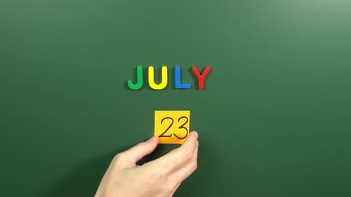 Hand sticking a sticker 23 July calendar day on school board. 23 date of July. Twenty third day