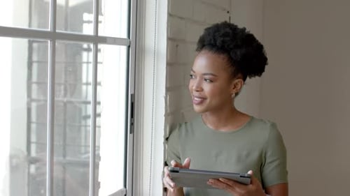 A young African American woman holds a tablet near a window, with copy space